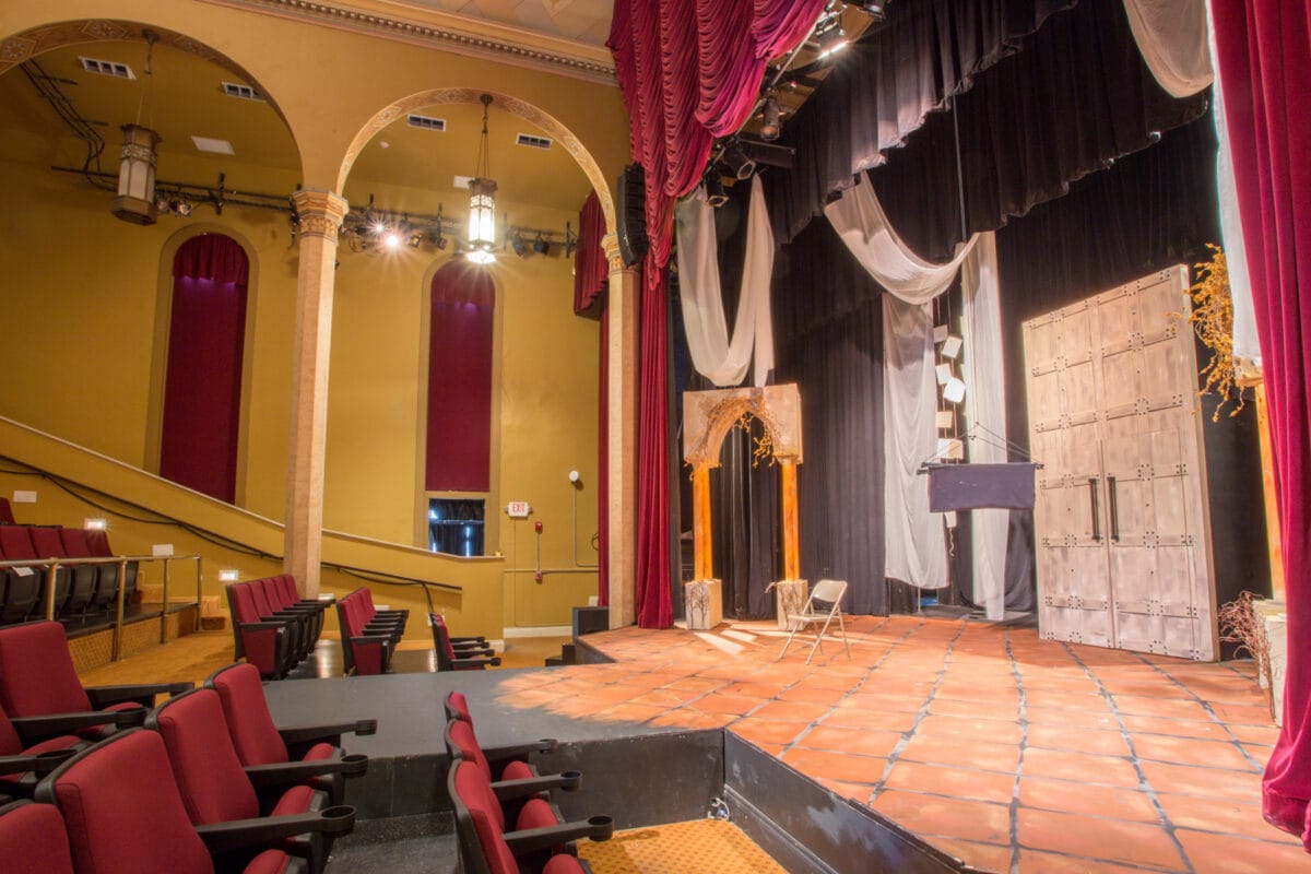 Empty theater stage with red seats, dramatic curtains, and set pieces under warm lighting.