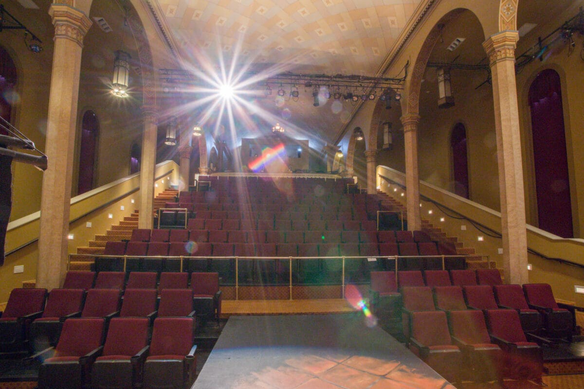 Rows of empty theater seats face a brightly lit stage in an ornate auditorium with arched walls.