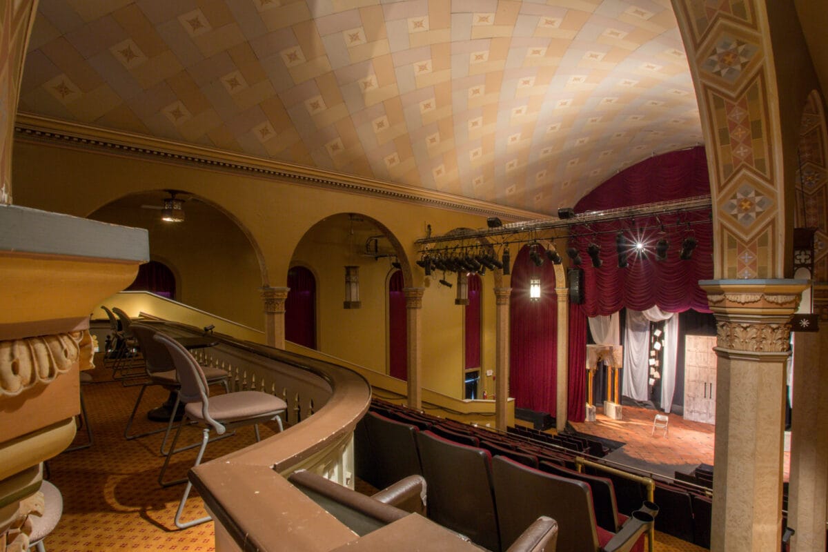 View of an ornate, empty theater with arched balcony seating and a stage with spotlights and red curtains.