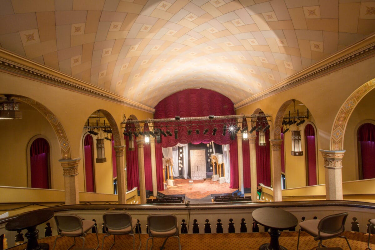 View of an empty, ornate theater with red curtains, arched columns, and stage from the balcony seats.