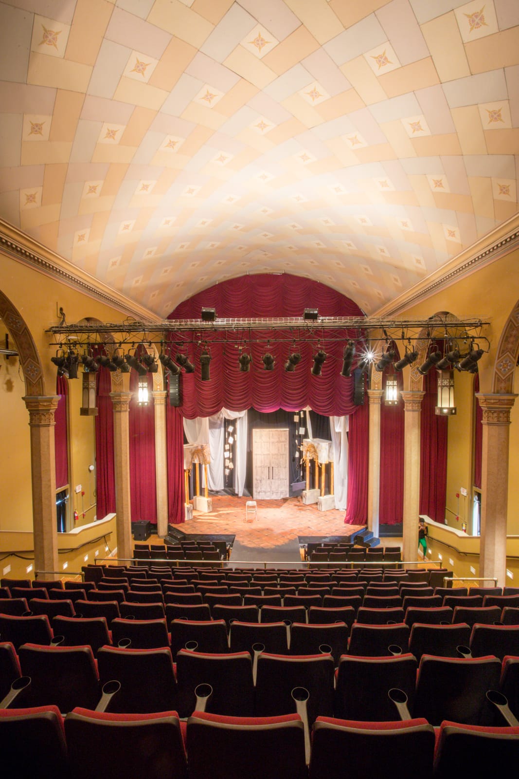 Empty theater with red seats facing a stage set for a performance, under a decorative arched ceiling.