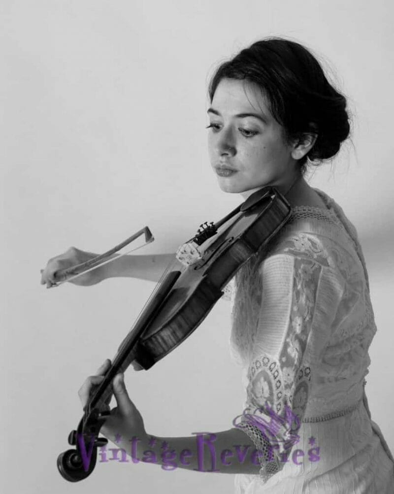 A black and white photo of a woman playing a violin in an Edwardian style outfit.