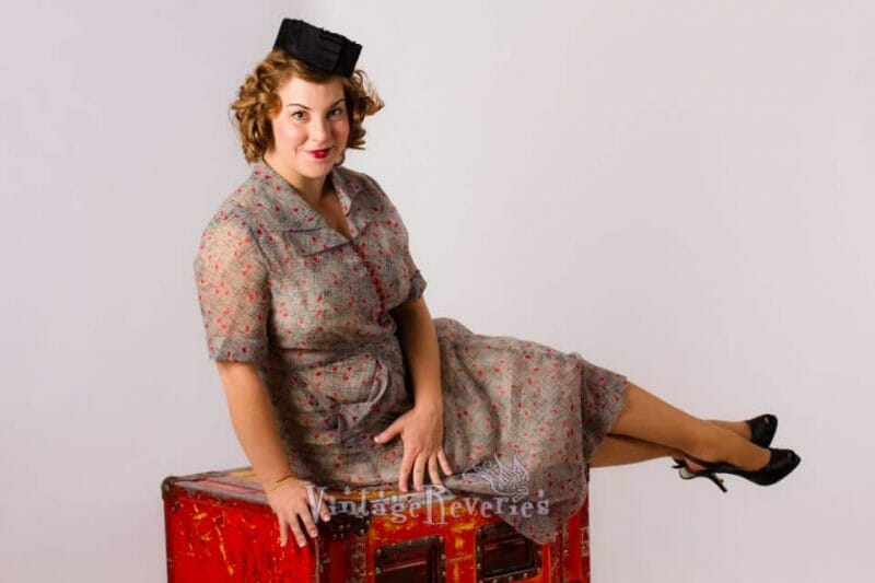 A woman in a vintage dress, channeling 1940s style, posing on top of a suitcase.