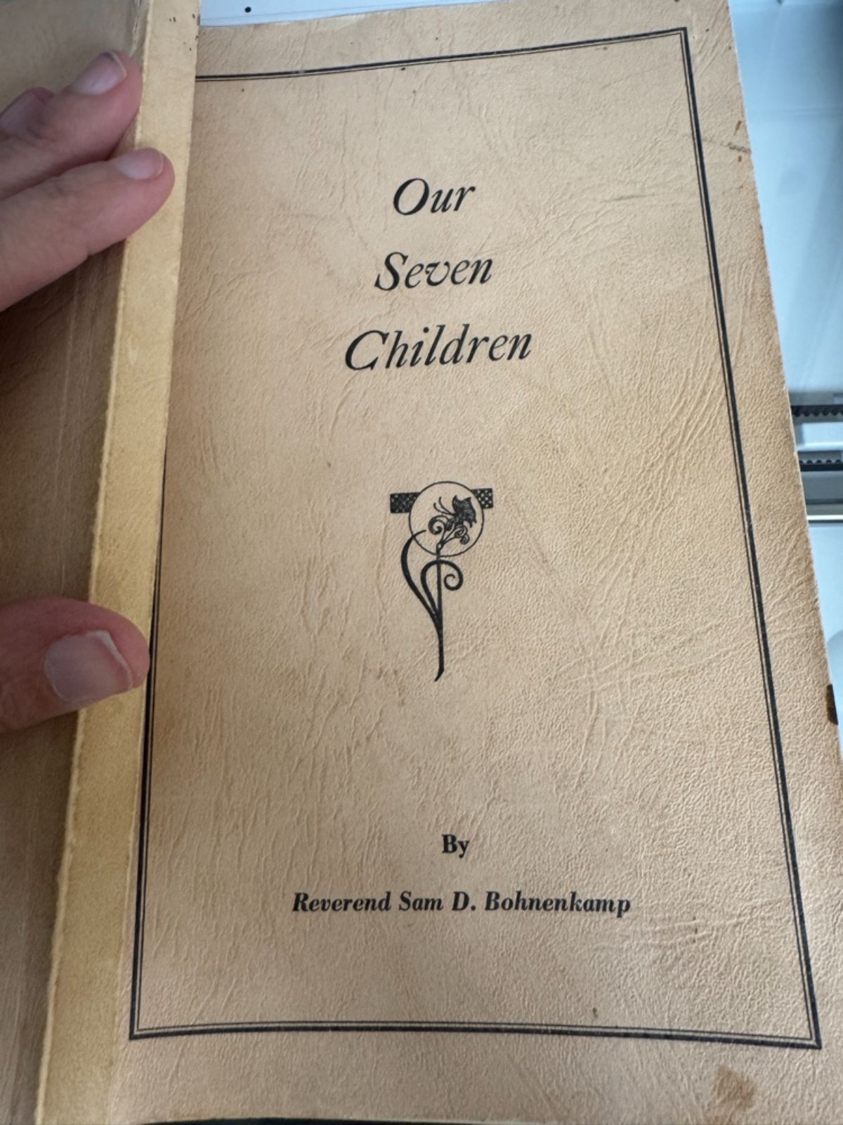Hand holding a vintage beige linen book with floral motif, titled “Our Seven Children” by Reverend Sam D. Bohnenkamp.
