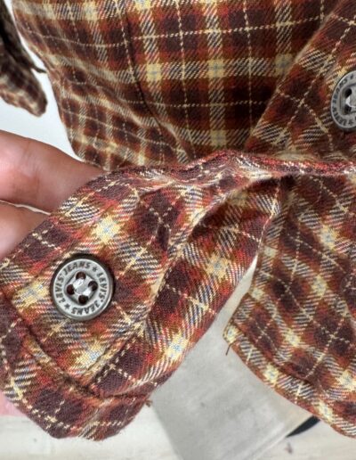 Vintage 90s Levi's brown plaid flannel shirt cuff, silver JEANS LEAVES buttons, visible stitching, classic grunge style.