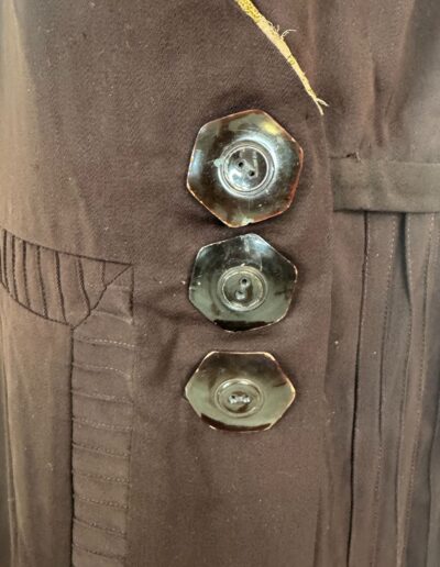 Close-up of a rare plus size 1920s dark brown drop waist dress with pleated details, hexagonal buttons, and loose thread at top seam.