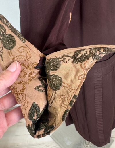 Hand holds brown and green embroidered fabric accessory beside rare plus size 1920s dark brown Art Deco drop waist dress XL/1X.