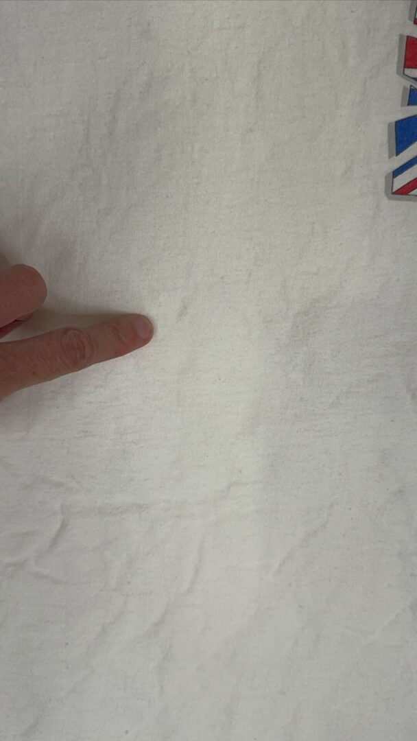 Finger points to a faint spot on white cotton vintage Def Leppard T-shirt; colorful graphic shapes in top right corner.
