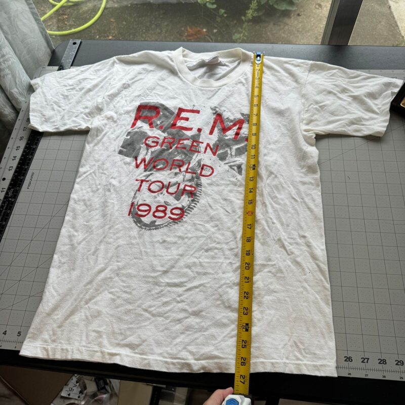 Vintage white 1989 R.E.M. tour T-shirt on a cutting mat with yellow measuring tape; window and greenery in background.