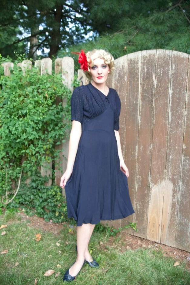 A woman in a Dainty Blue 1930s Vintage Dress stands in front of a fence.